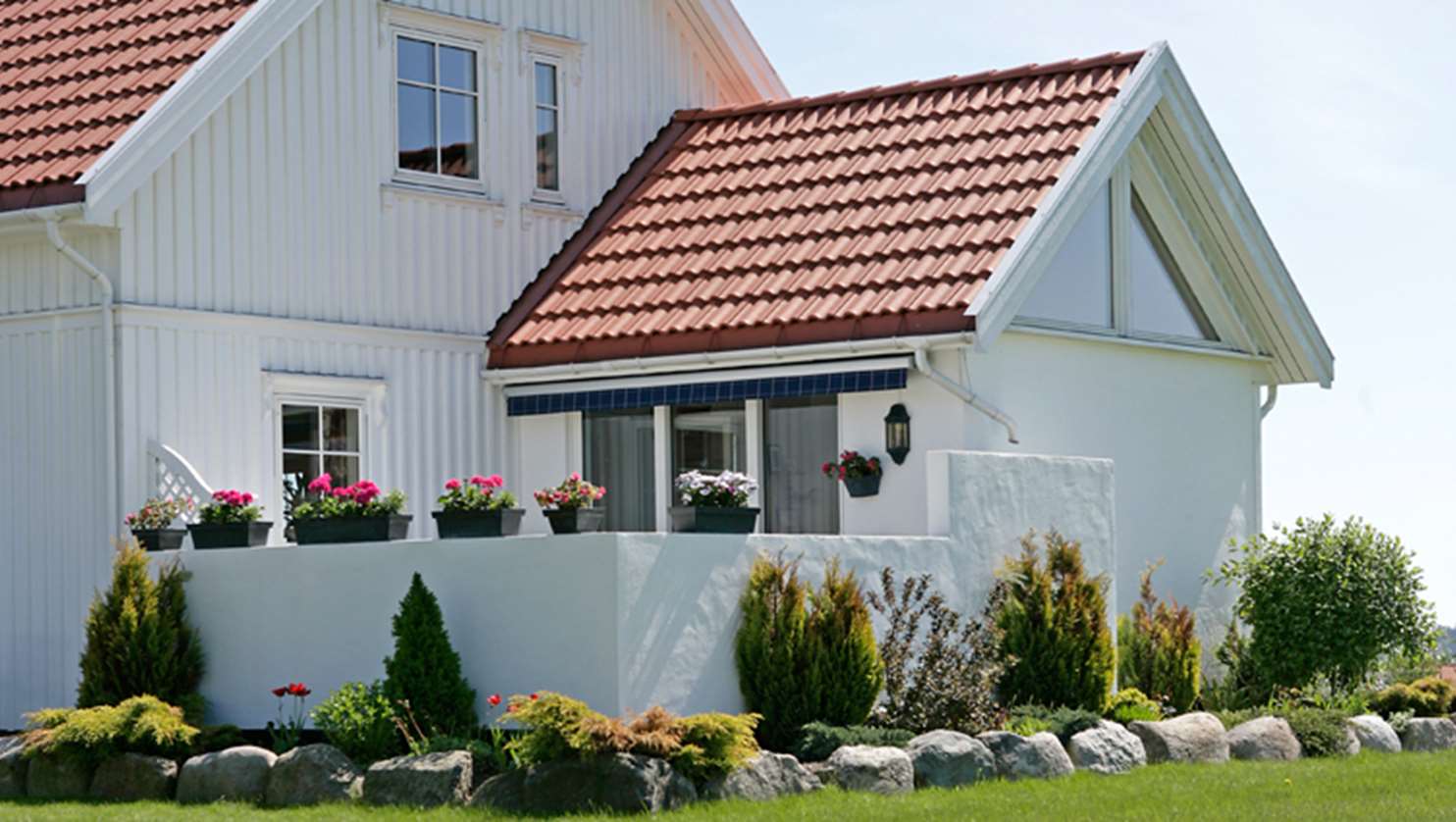 Villa med blommande trädgård. Vitt hus med tegelröda takpannor i betong.