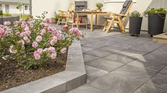 Trädgårdsmiljö med plattor, kantsten och blommor framför ett hus.